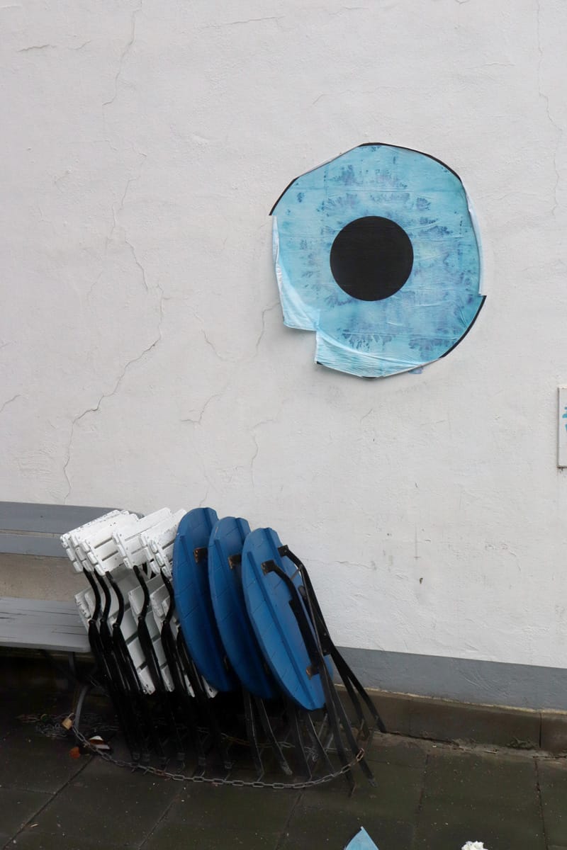 Blaues Papierauge an heller Mauerwand. Davor zusammengeklappt Bistrostühle aus Holz